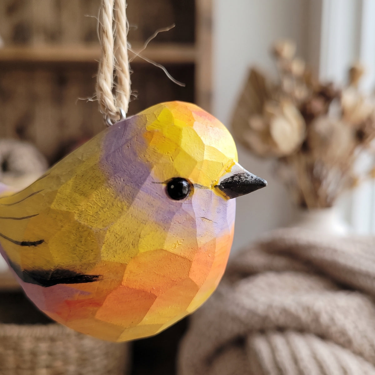 A faceted wooden White-browed tit-warbler hanging ornament painted in yellow, orange, and purple tones, with a black beak and eye, hanging by a twine string in a cozy indoor setting.