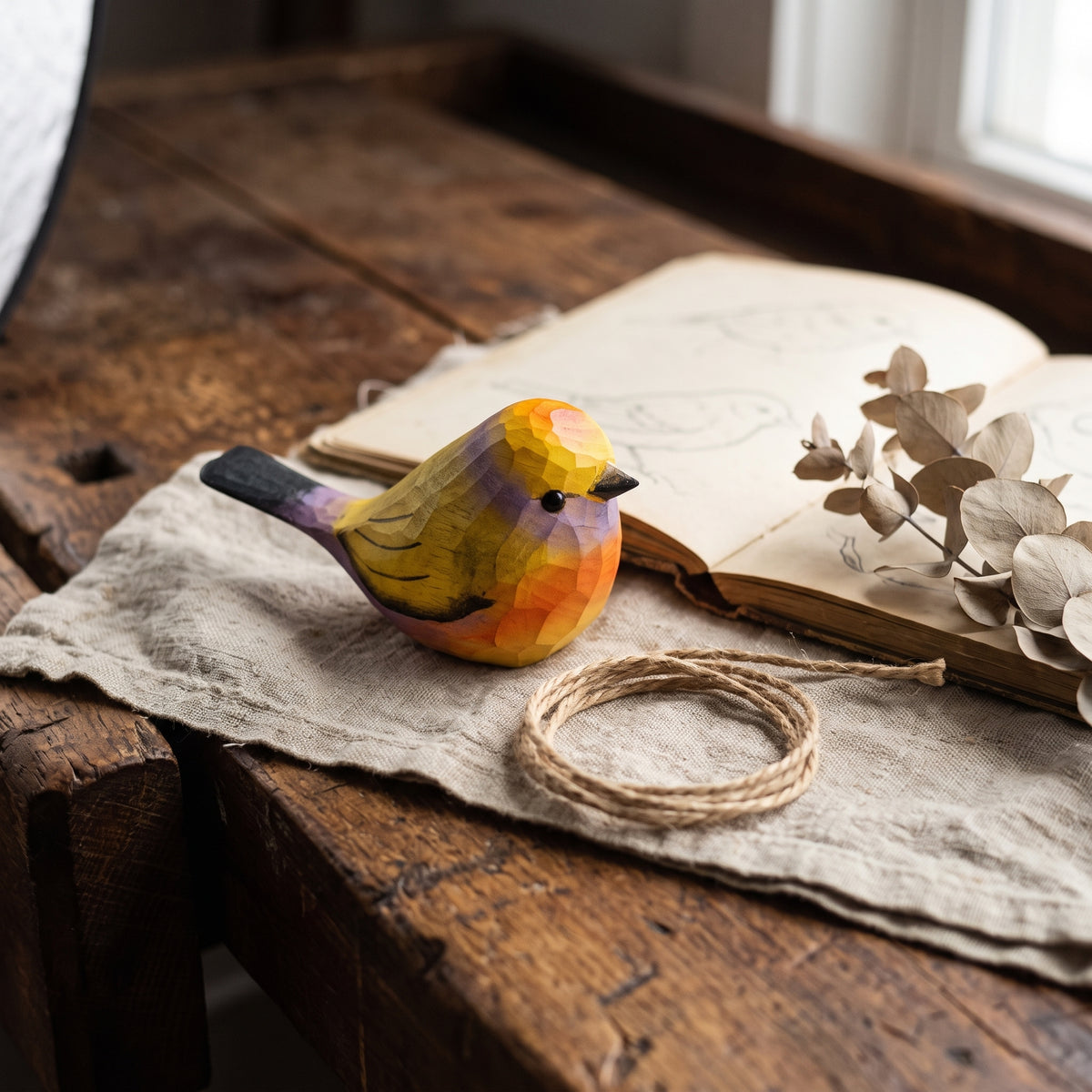 A small hand-painted wooden White-browed tit-warbler figurine with a geometric, faceted low-poly design, resting on a rustic wooden table next to an open book, dried eucalyptus leaves, and a coiled piece of twine.