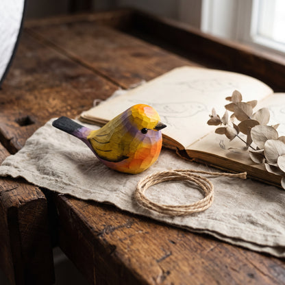 A small hand-painted wooden White-browed tit-warbler figurine with a geometric, faceted low-poly design, resting on a rustic wooden table next to an open book, dried eucalyptus leaves, and a coiled piece of twine.