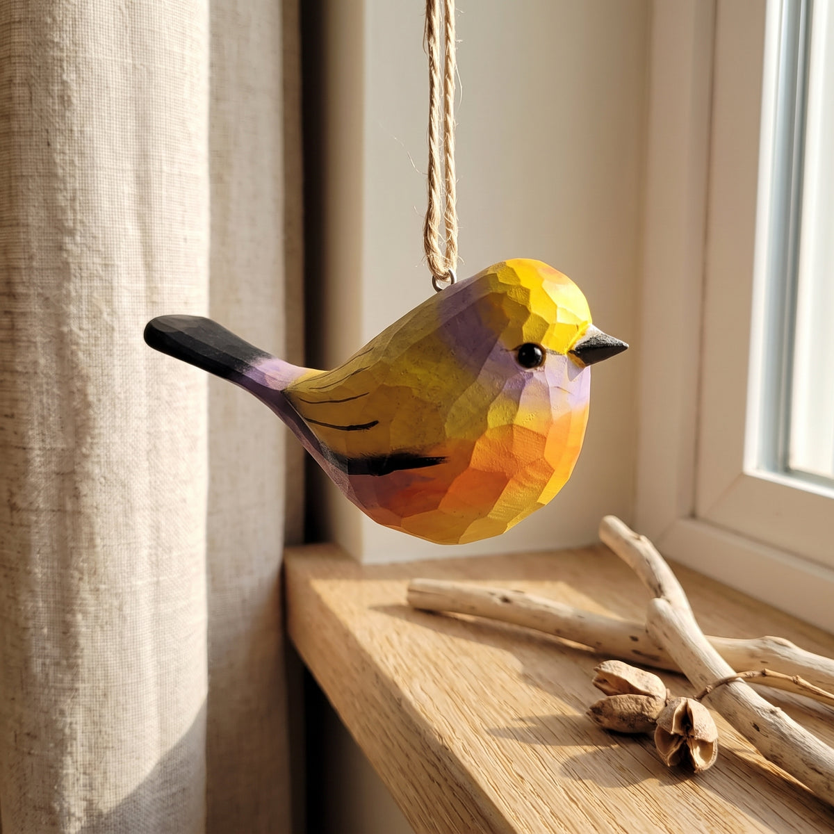A faceted wooden White-browed tit-warbler ornament in yellow, orange, and purple hangs from a rope near a window on a wooden windowsill, with driftwood and a curtain in the background.