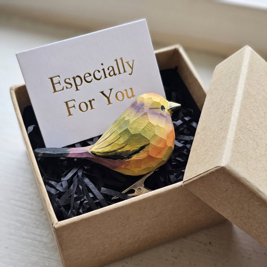 A hand-carved wooden White-Browed Tit-Warbler clip-on ornament resting in an open kraft gift box filled with black shredded paper, featuring a yellow and soft lavender-tinted body with black wing accents, and a small card reading "Especially For You" placed behind it.