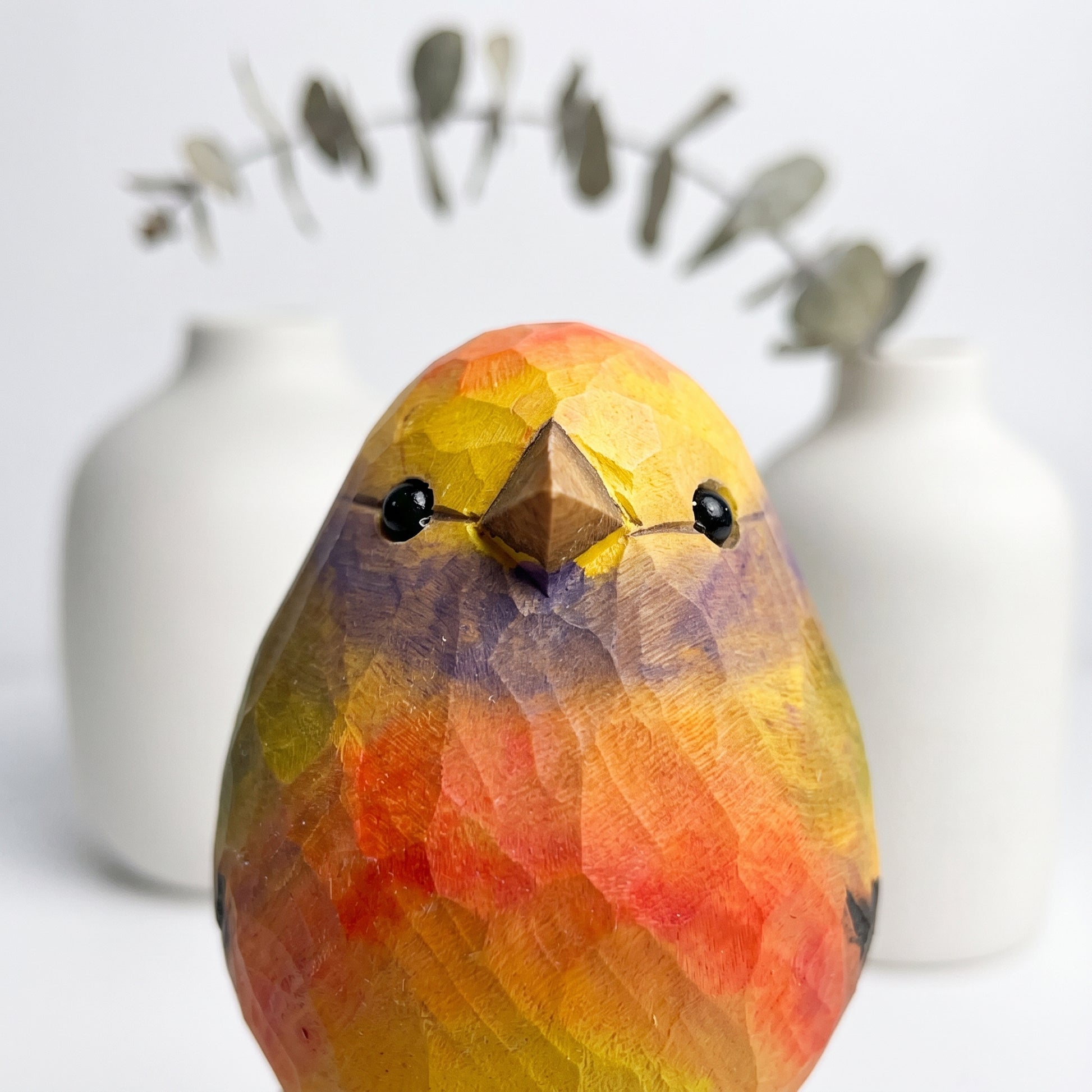 A carved wooden White-browed tit-warbler sculpture with a faceted, geometric texture, painted in vibrant yellow, orange, and red with a brown beak and black eyes. It is photographed in a studio setting with two blurred white ceramic vases in the background.