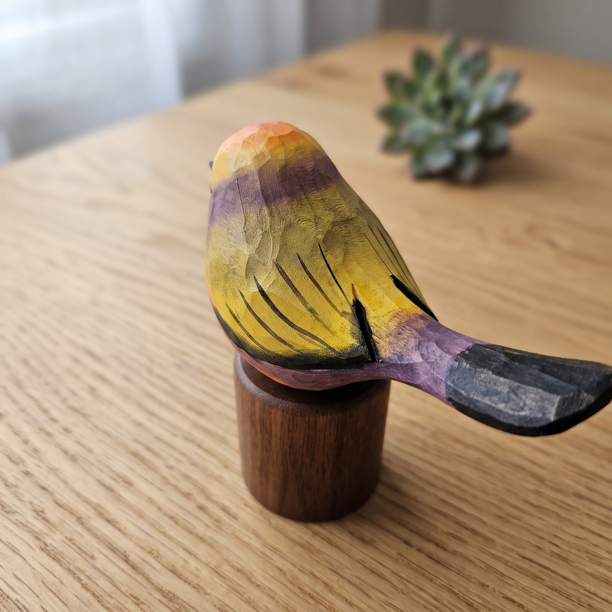 A hand-carved wooden White-browed tit-warbler figurine painted with yellow, purple, black, and orange, mounted on a small wooden base on a light wood tabletop.
