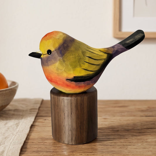 A hand-painted wooden White-browed tit-warbler figurine with yellow, green, orange, and black feathers, mounted on a dark wooden cylinder pedestal, displayed on a rustic wooden table next to a bowl of fruit.