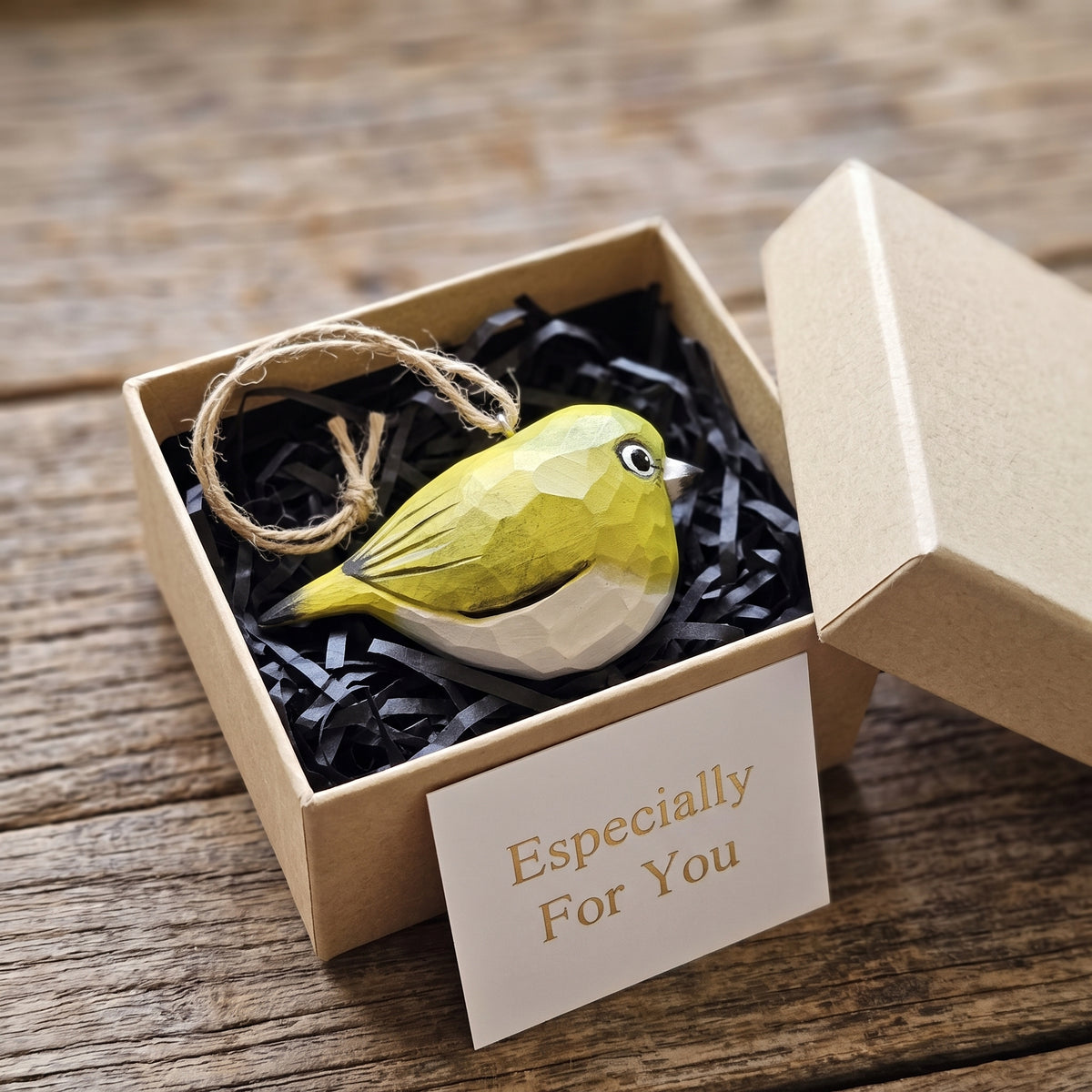 A small White-Eyes-shaped ornament in yellow-green and white sits in black shredded paper inside an open kraft gift box, with a card reading 'Especially For You' on the front and a rustic wood surface around it.