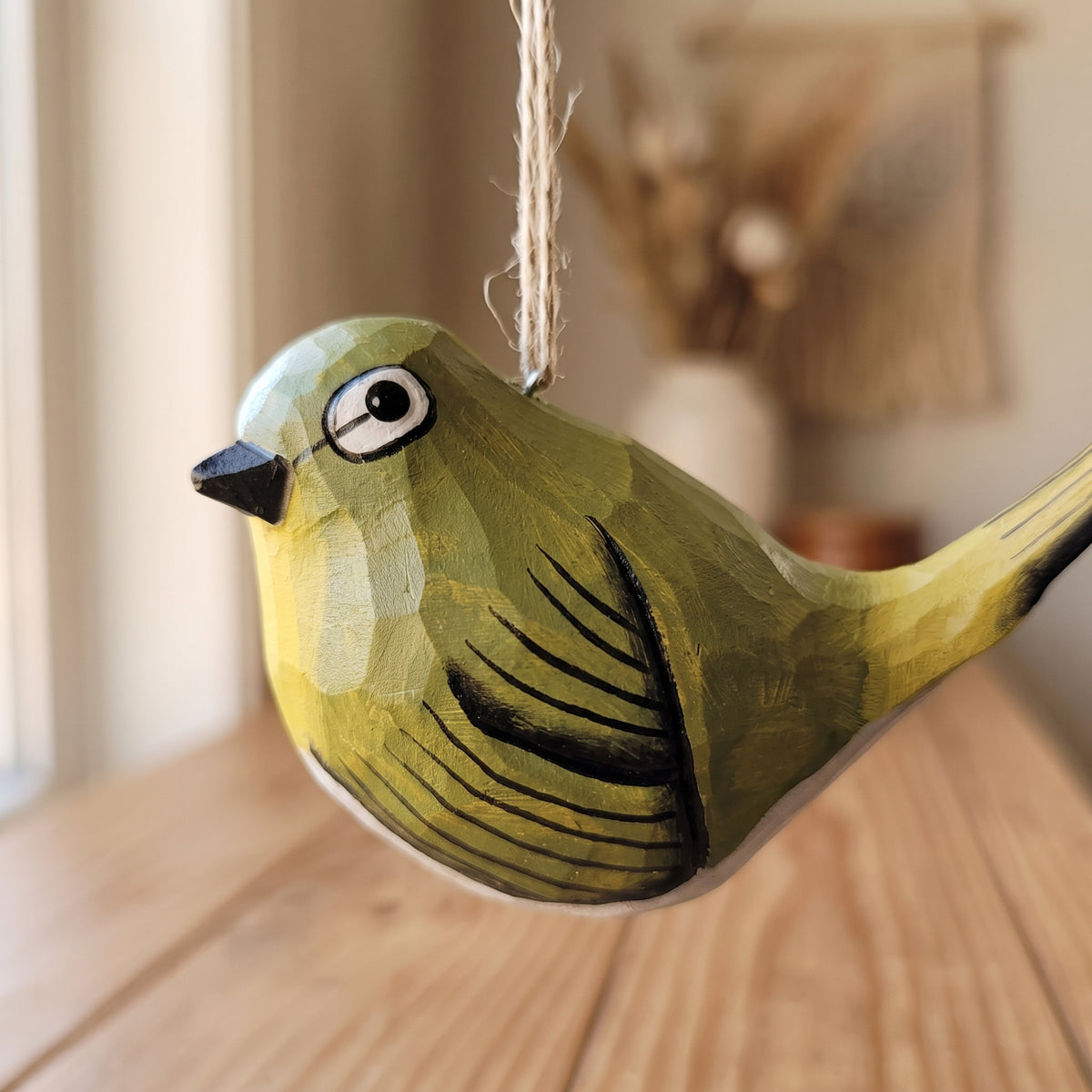 A hand-painted wooden White-Eyes ornament in shades of olive green and yellow, hanging by a twine string in a warm, softly blurred indoor setting.