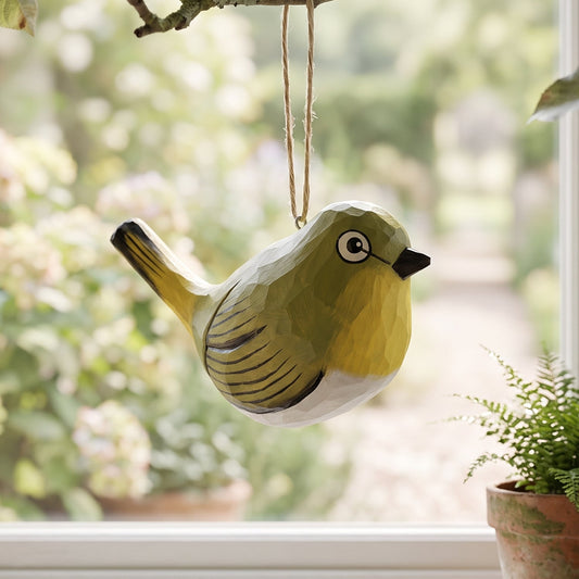 A small hanging White-Eyes ornament with olive green, yellow, and white coloring hangs from a tree branch in front of a window. A potted fern sits on the windowsill, and blurred garden foliage fills the background.
