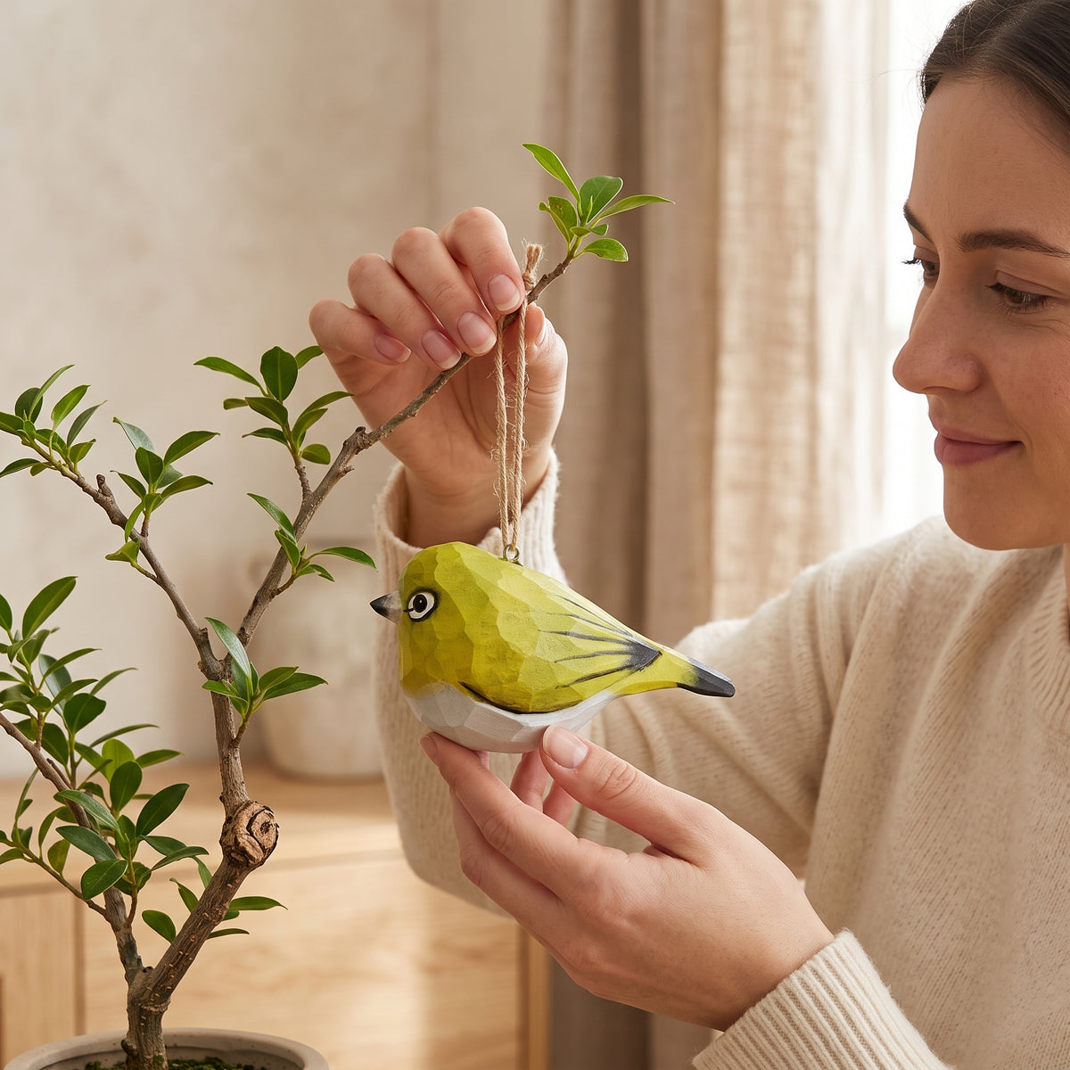 A woman holds a small green and White-Eyes bird-shaped hanging ornament with a string near a potted plant in a softly lit indoor setting.