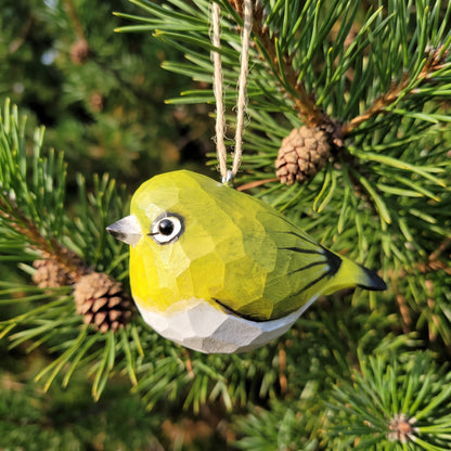 A small White-Eyes-shaped hanging ornament shaped like a green bird with a white belly, hanging from a string among pine branches and pinecones.