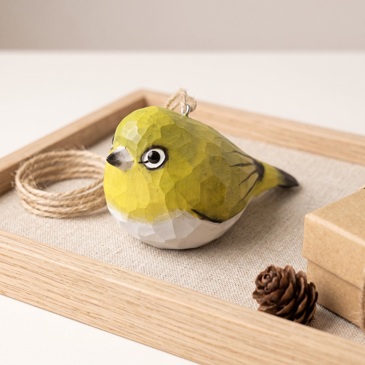 A small White-Eyes-shaped ornament with a faceted geometric texture, painted in olive green and white with black details, resting on a wooden tray with twine, a pinecone, and a box.