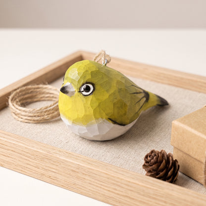 A small White-Eyes-shaped ornament with a faceted geometric texture, painted in olive green and white with black details, resting on a wooden tray with twine, a pinecone, and a box.