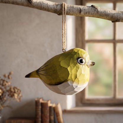 A geometric, faceted White-Eyes ornament in olive green, yellow, and white hangs from a natural twine loop attached to a birch branch, set against a softly blurred indoor background with a window and books.