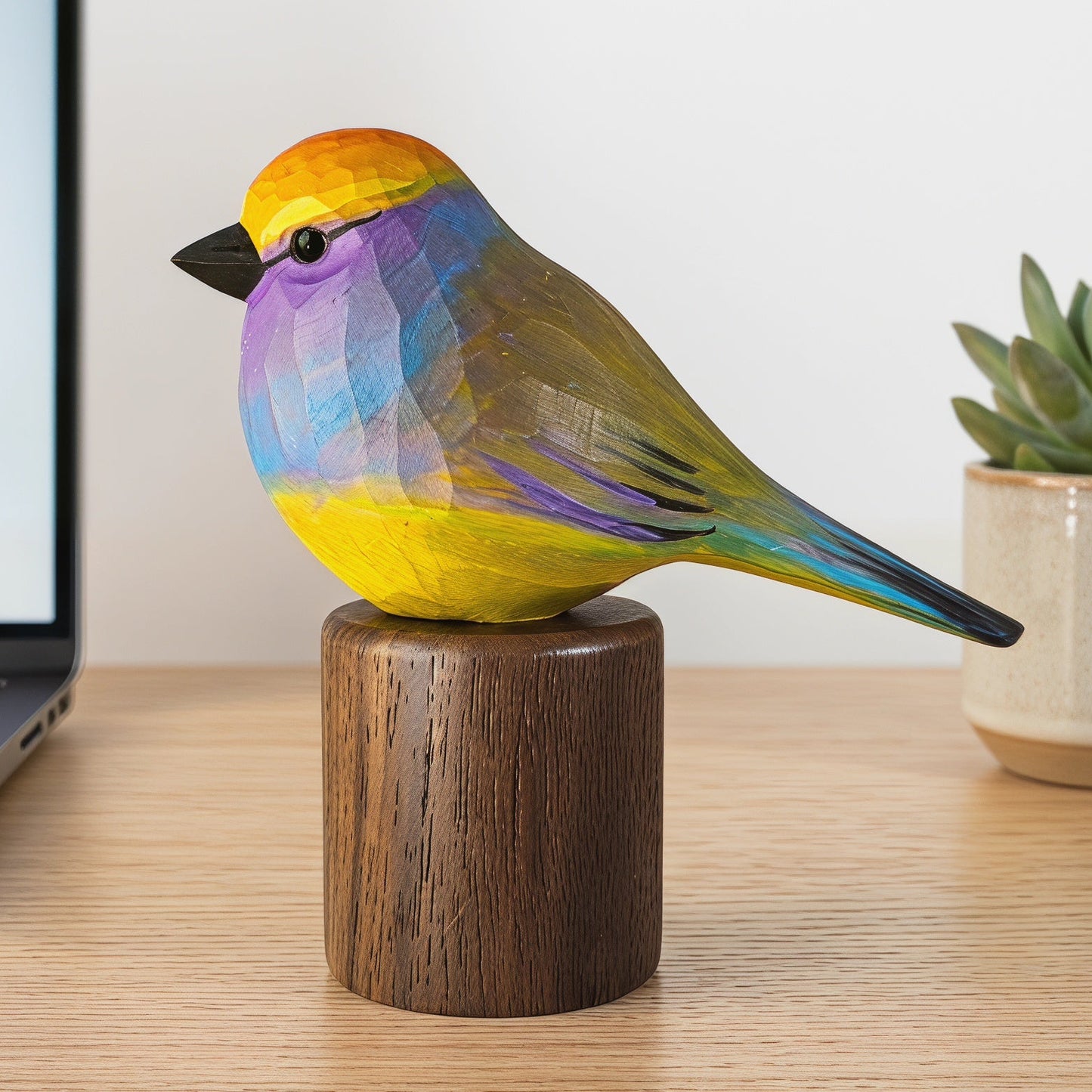 Hand-carved wooden White-browed Tit-warbler figurine painted in yellow, blue, and purple tones, perched on a cylindrical black walnut base on an office desk next to a laptop and a potted succulent.