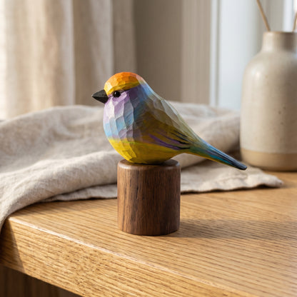 Hand-carved wooden White-browed Tit-warbler bird figurine with colorful yellow, blue, and violet plumage, perched on a cylindrical black walnut base on a wooden tabletop next to a folded cloth and ceramic vase.