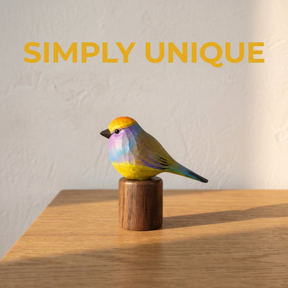 Promotional graphic with the text "SIMPLY UNIQUE" featuring a hand-carved wooden White-browed Tit-warbler figurine in vibrant yellow, violet, and blue perched on a cylindrical black walnut base on a sunlit wooden table.