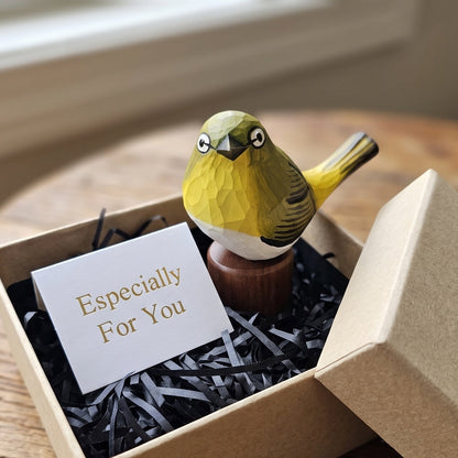 A small hand-painted White-eyes figurine in yellow, green, and white sits in a kraft gift box filled with black paper shred and a card that reads 'Especially For You.' The box is placed on a wooden table near a window.