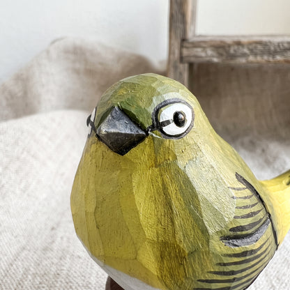 A close-up of a hand-carved wooden White-eyes figurine painted in olive green and yellow with black markings, set against a soft beige fabric background.