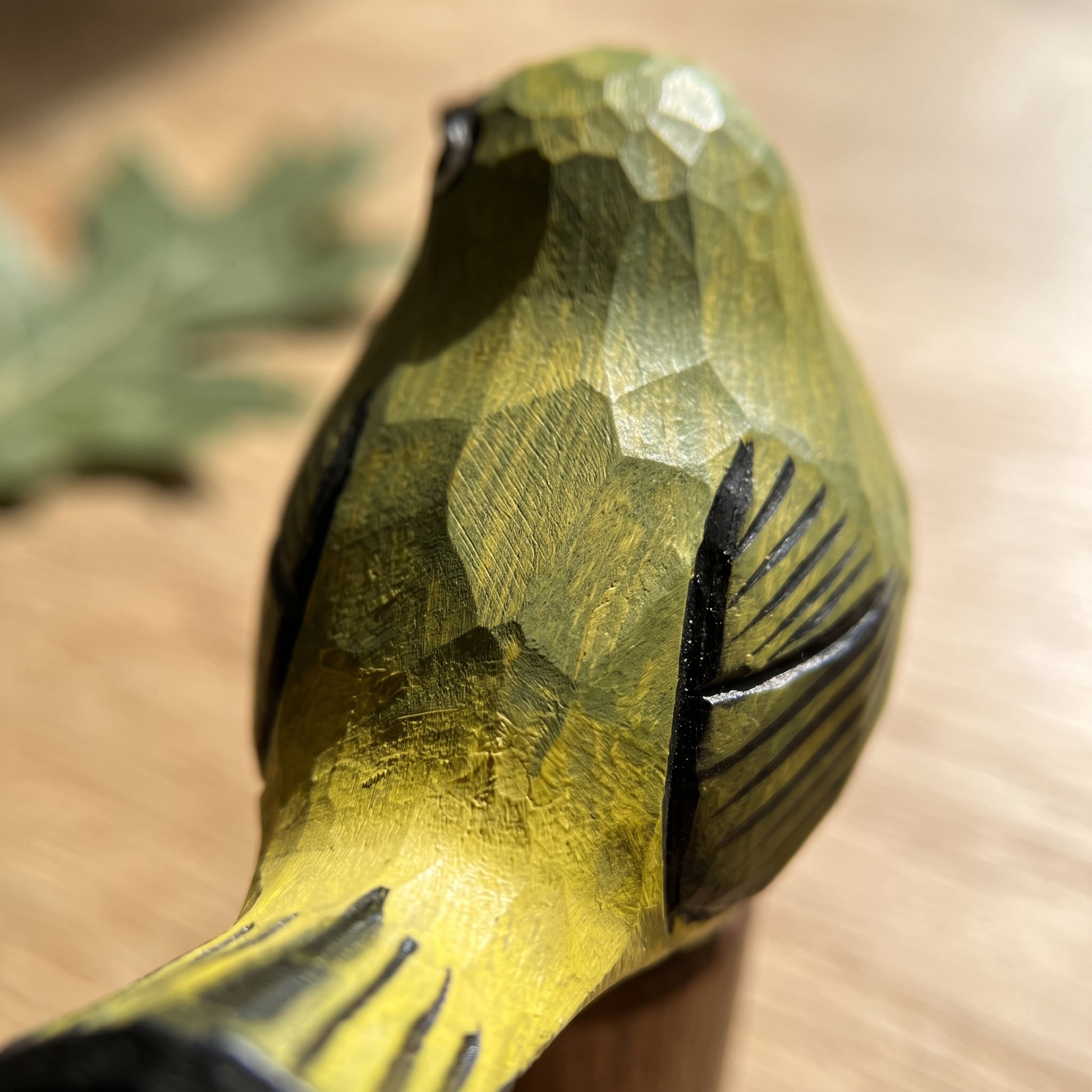 A close-up of a hand-carved wooden White-eyes figurine with a textured yellow-green and black finish, resting on a light wood surface.