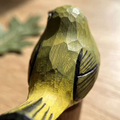 A close-up of a hand-carved wooden White-eyes figurine with a textured yellow-green and black finish, resting on a light wood surface.