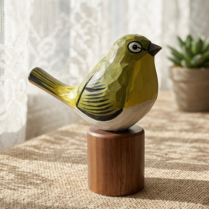 A small carved White-eyes bird figurine in olive green, cream, and black sits on a round wooden pedestal on a woven tablecloth, with a potted plant and lace curtain in the softly lit background.