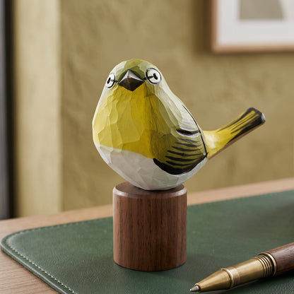 A small carved White-eyes figurine with a yellow-green, white, and black faceted geometric design, mounted on a round wooden pedestal. The bird sculpture is displayed on a desk next to a green leather pad, a pen, and a ceramic mug.