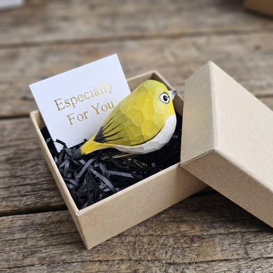 A hand-carved wooden White-Eye clip-on bird ornament resting inside an open kraft paper gift box filled with black shredded paper, featuring a vibrant yellow head and breast, a distinct white ring around its eye, and a white card reading "Especially For You" placed in the foreground on a rustic wooden surface.
