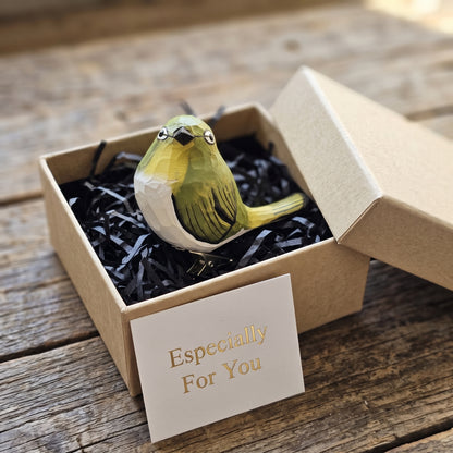 A hand-carved wooden White-Eye clip-on bird ornament resting inside an open kraft paper gift box filled with black shredded paper, featuring olive-green, cream, and yellow plumage with a distinct white ring around its eye, alongside a white card reading "Especially For You" in the foreground on a rustic wooden surface.