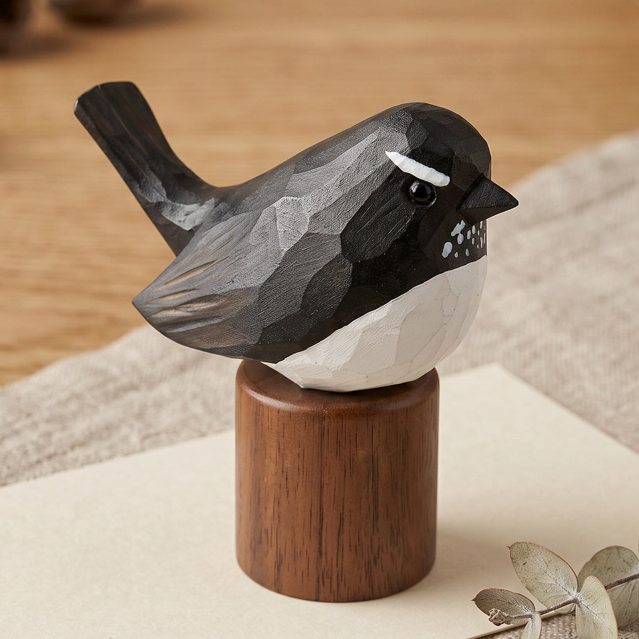 A carved wooden Willie wagtail figurine with a black and white body perched on a round brown wooden base, styled on a beige cloth over a light wood tabletop with dried acorns and a ceramic bowl in the background.