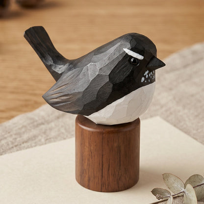 A carved wooden Willie wagtail figurine with a black and white body perched on a round brown wooden base, styled on a beige cloth over a light wood tabletop with dried acorns and a ceramic bowl in the background.