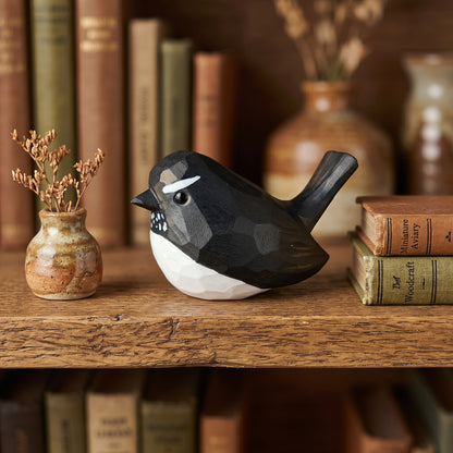 A small Willie wagtail-shaped decorative object with a black and white geometric finish sits on a wooden shelf beside a tiny vase with dried stems and stacked vintage books.