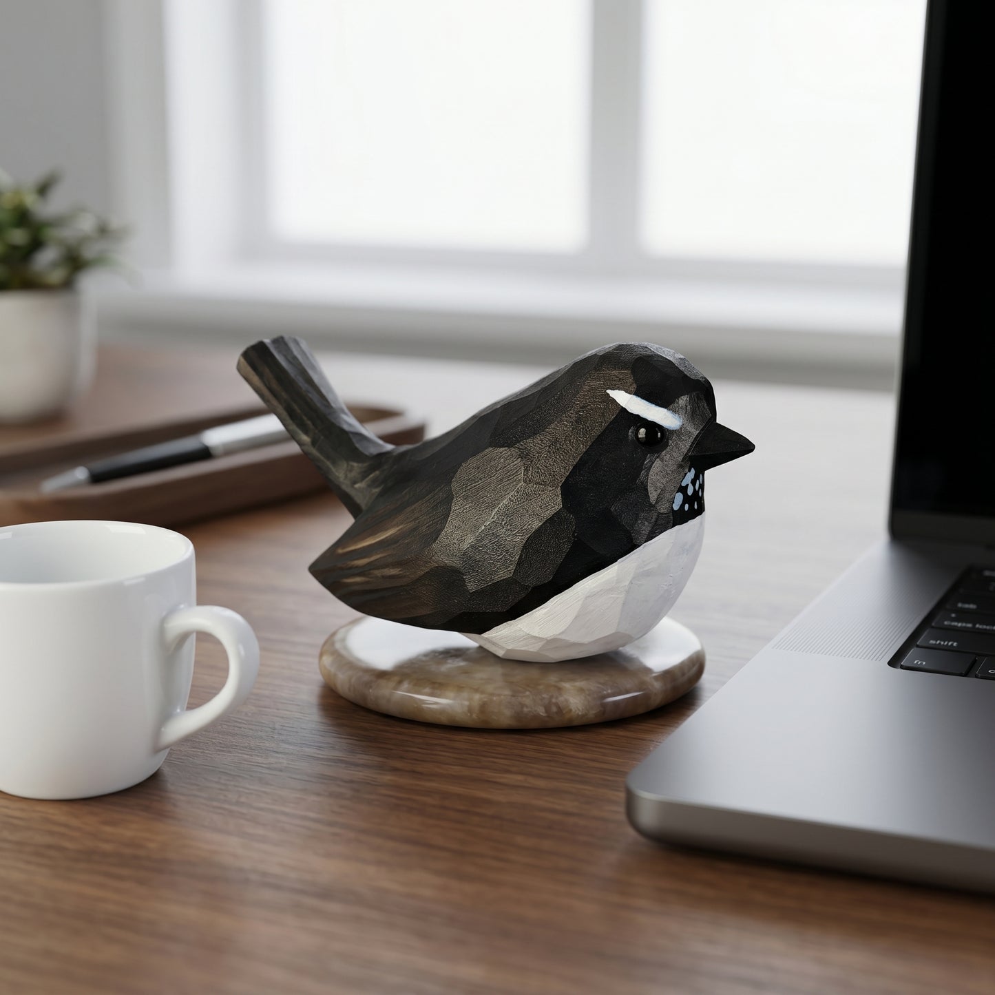 Hand-carved wooden Willie Wagtail figurine with black and white plumage sitting on a desk next to a laptop and coffee mug.