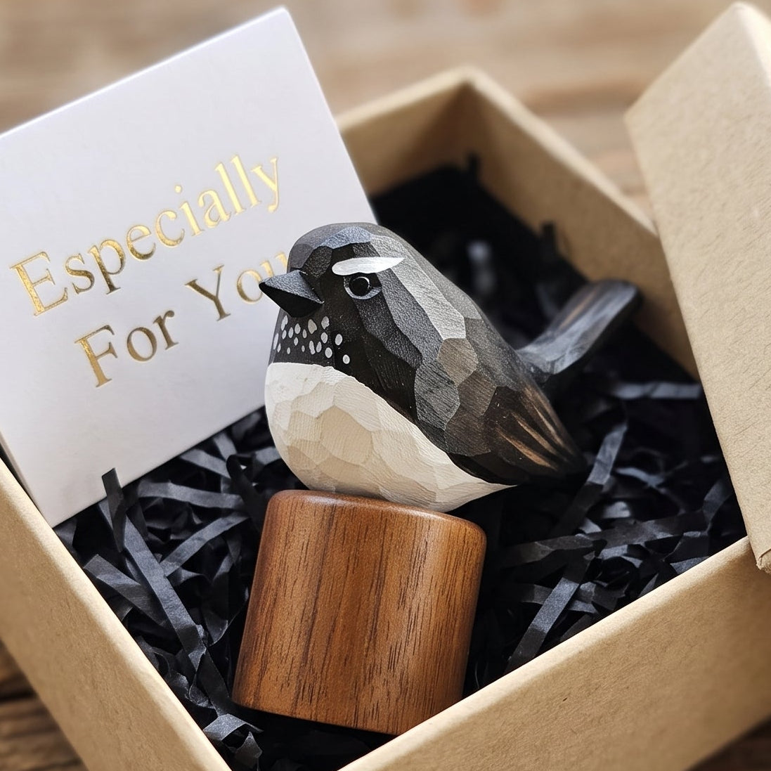 A small carved Willie wagtail figurine resting on a wooden base inside an open kraft gift box with black shredded paper filler and a card that reads 'Especially For You,' placed on a rustic wooden surface.
