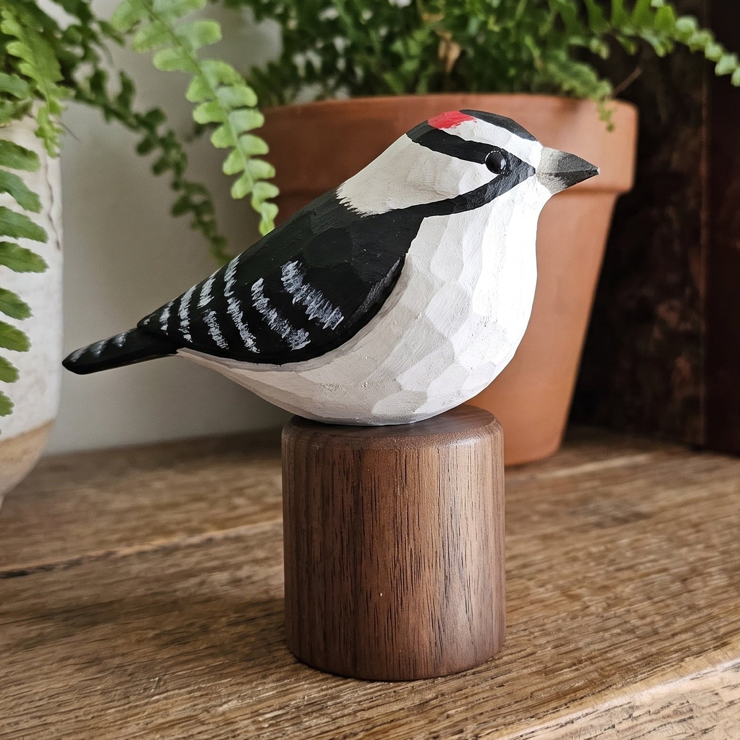 A carved wooden Downy Woodpecker figurine with black, white, and red painted details sits on a small cylindrical wooden base on a rustic wooden shelf, with a potted fern and old books in the background.