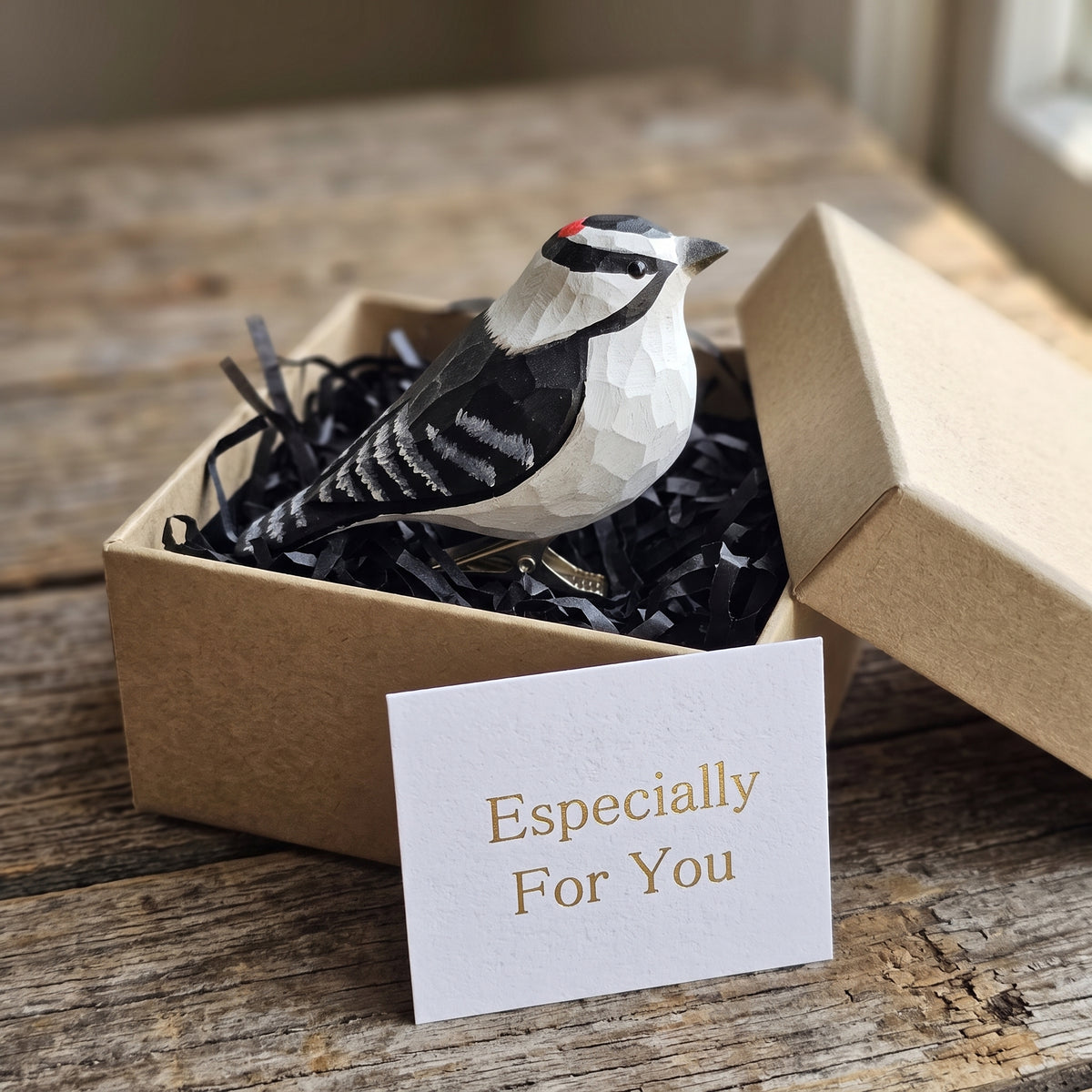 Hand-carved wooden Downy Woodpecker clip-on ornament resting in black shredded paper inside an open kraft gift box, with a white gift card reading "Especially For You" in front.