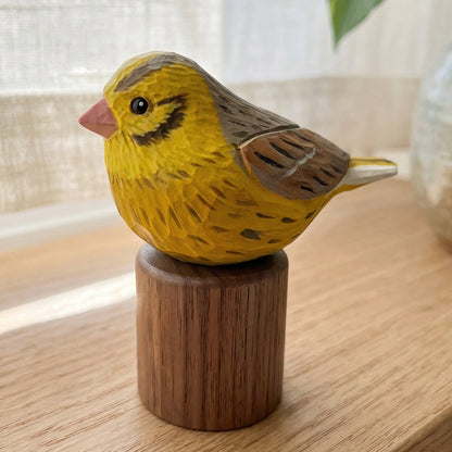 A hand-carved wooden Yellowhammer figurine, resembling a yellow canary with brown wings and a pink beak, perched on a cylindrical wooden base. The figurine is displayed on a light wood table in a softly lit room with a window and a ceramic vase in the background.