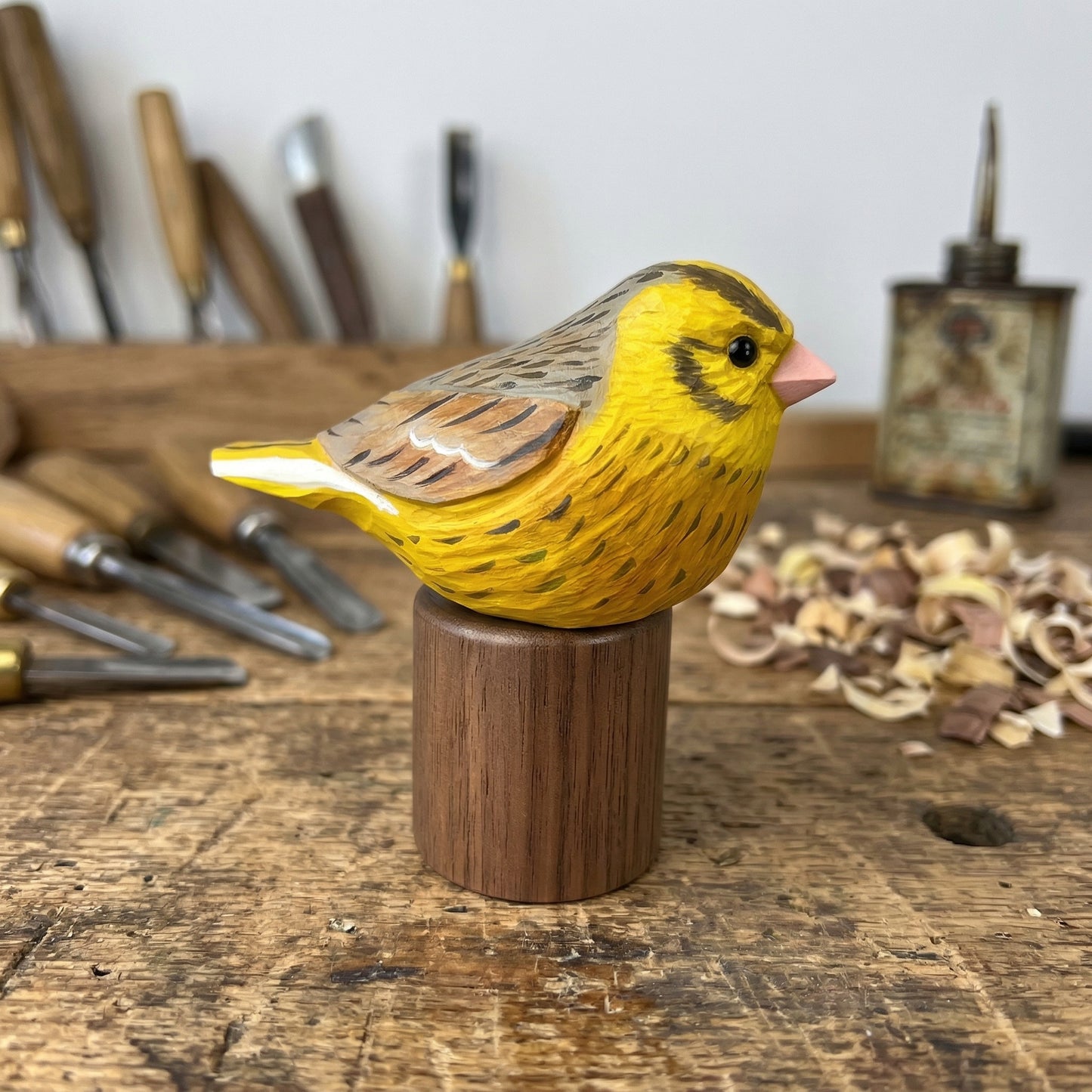 A hand-carved wooden Yellowhammer figurine, resembling a canary, painted bright yellow with brown and black markings, perched on a cylindrical wooden base on a rustic workbench with carving tools and wood shavings in the background.