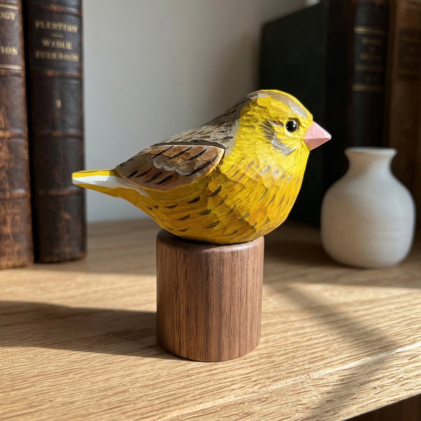 A carved wooden Yellowhammer figurine, likely a canary, painted bright yellow with brown wings and a pink beak, mounted on a cylindrical wooden base on a wooden shelf.