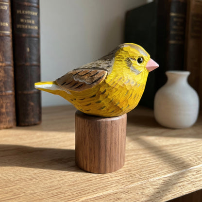 A carved wooden Yellowhammer figurine, likely a canary, painted bright yellow with brown wings and a pink beak, mounted on a cylindrical wooden base on a wooden shelf.