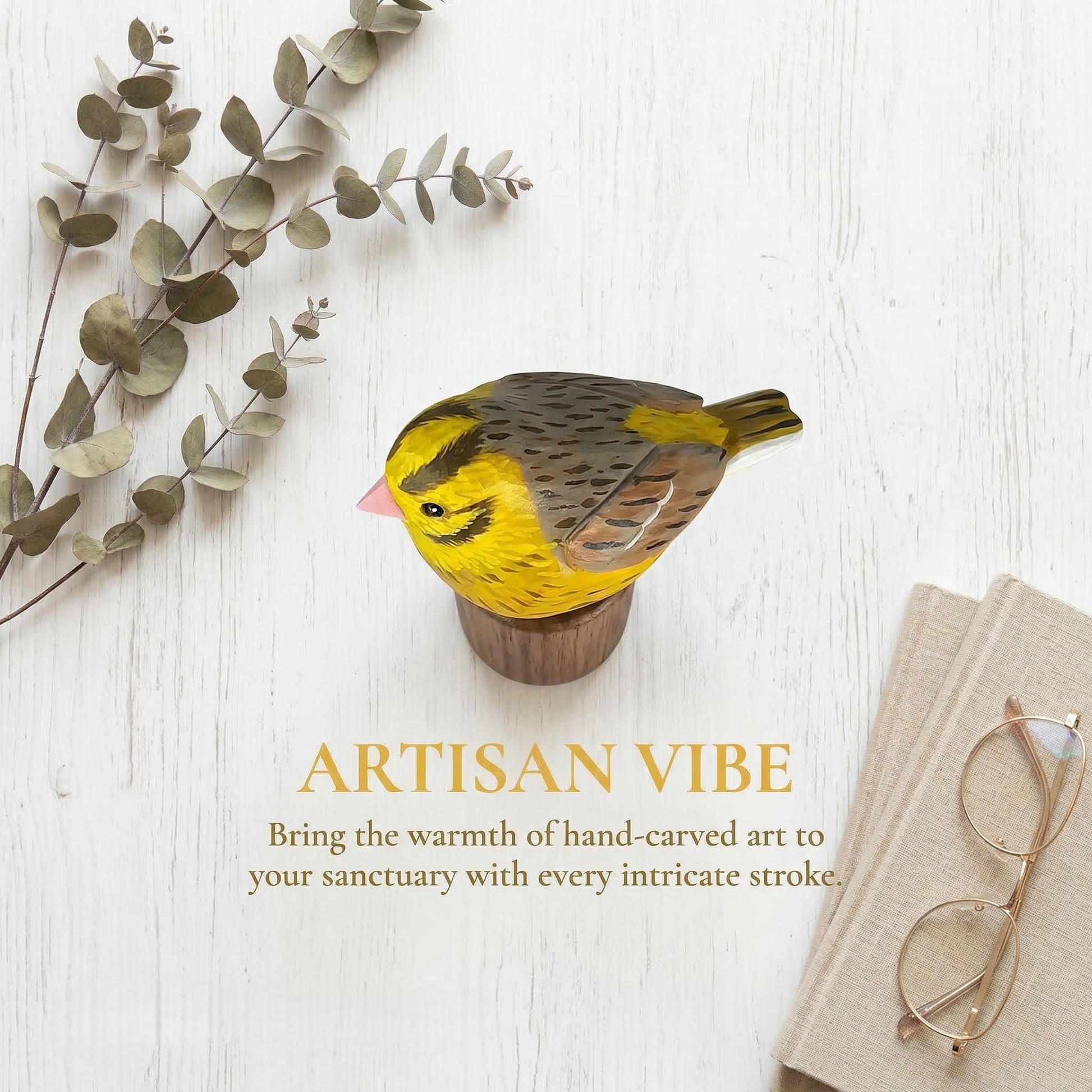 A hand-carved wooden Yellowhammer figurine with a bright yellow chest, brown and gray wings, and a pink beak, displayed on a small wooden base. The scene is styled on a white wood background with eucalyptus branches, folded beige fabric, and eyeglasses, with the text 'ARTISAN VIBE' and a subtitle below.