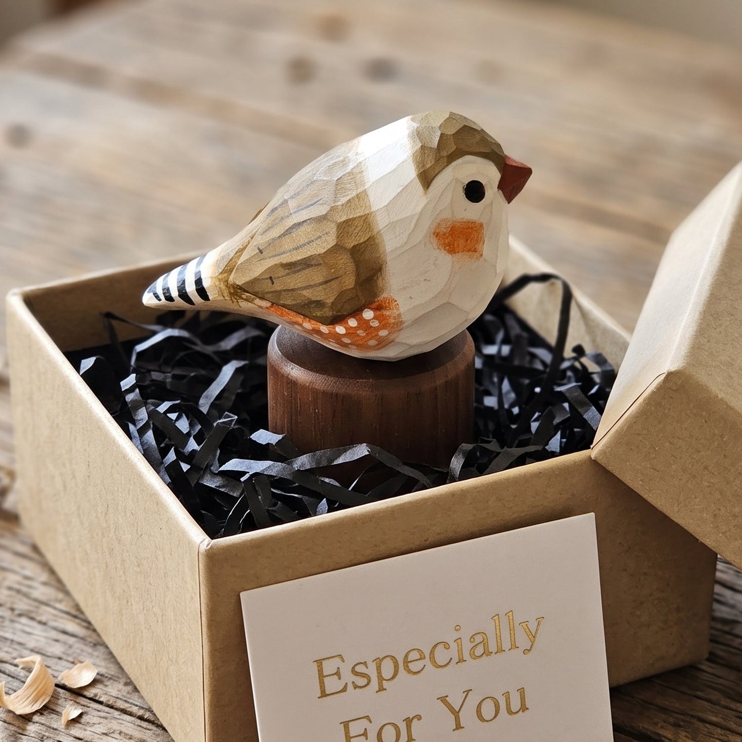 A small carved wooden Zebra Finch figurine sits in a kraft gift box filled with black shredded paper, with a card reading 'Especially For You' in front. The scene is styled on a rustic wooden tabletop.
