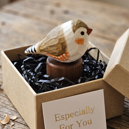 A small carved wooden Zebra Finch figurine sits in a kraft gift box filled with black shredded paper, with a card reading 'Especially For You' in front. The scene is styled on a rustic wooden tabletop.
