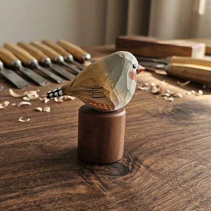 A hand-carved wooden Zebra Finch figurine with geometric facets, perched on a cylindrical wooden base, set on a rustic wooden table with woodworking tools in the background.