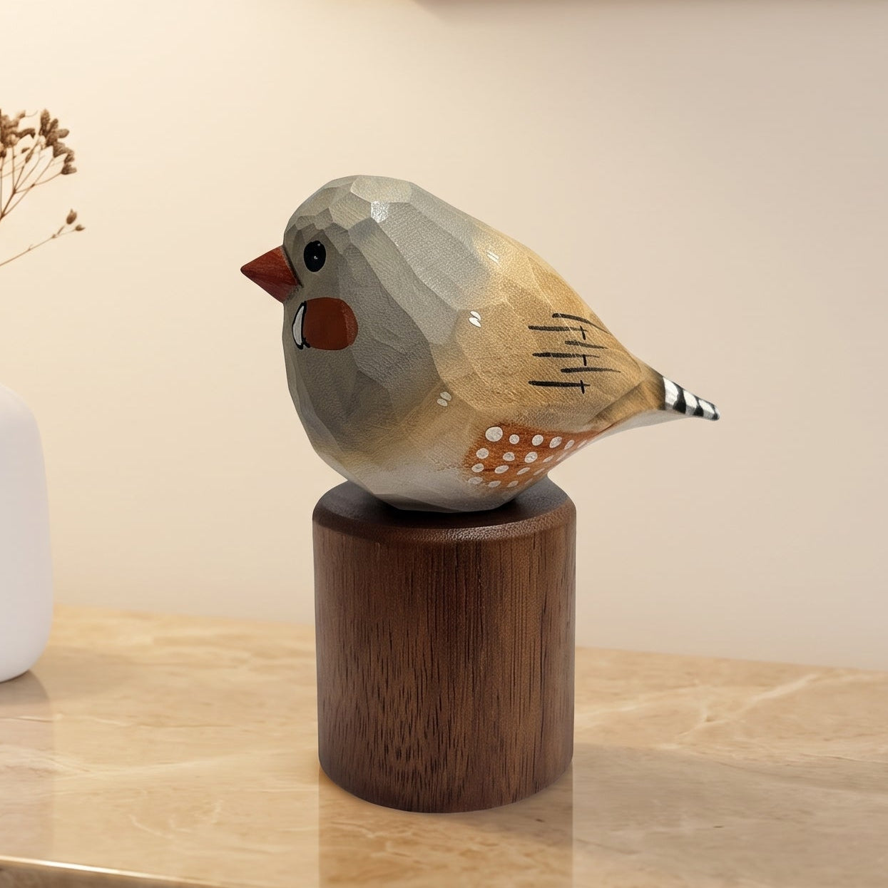 A decorative Zebra Finch figurine with a geometric, faceted finish in muted gray, beige, and orange-brown tones, perched on a dark wooden cylindrical base. The figurine is styled on a beige marble side table next to a white ceramic vase with dried stems, with a warm lamp and framed wall art in the background.