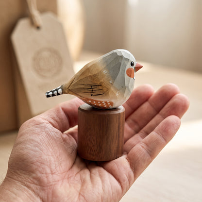 A hand holds a small carved wooden Zebra Finch ornament on a cylindrical wooden base. The bird has a white head, gray wings, a peach beak, and brown and white spotted feathers.
