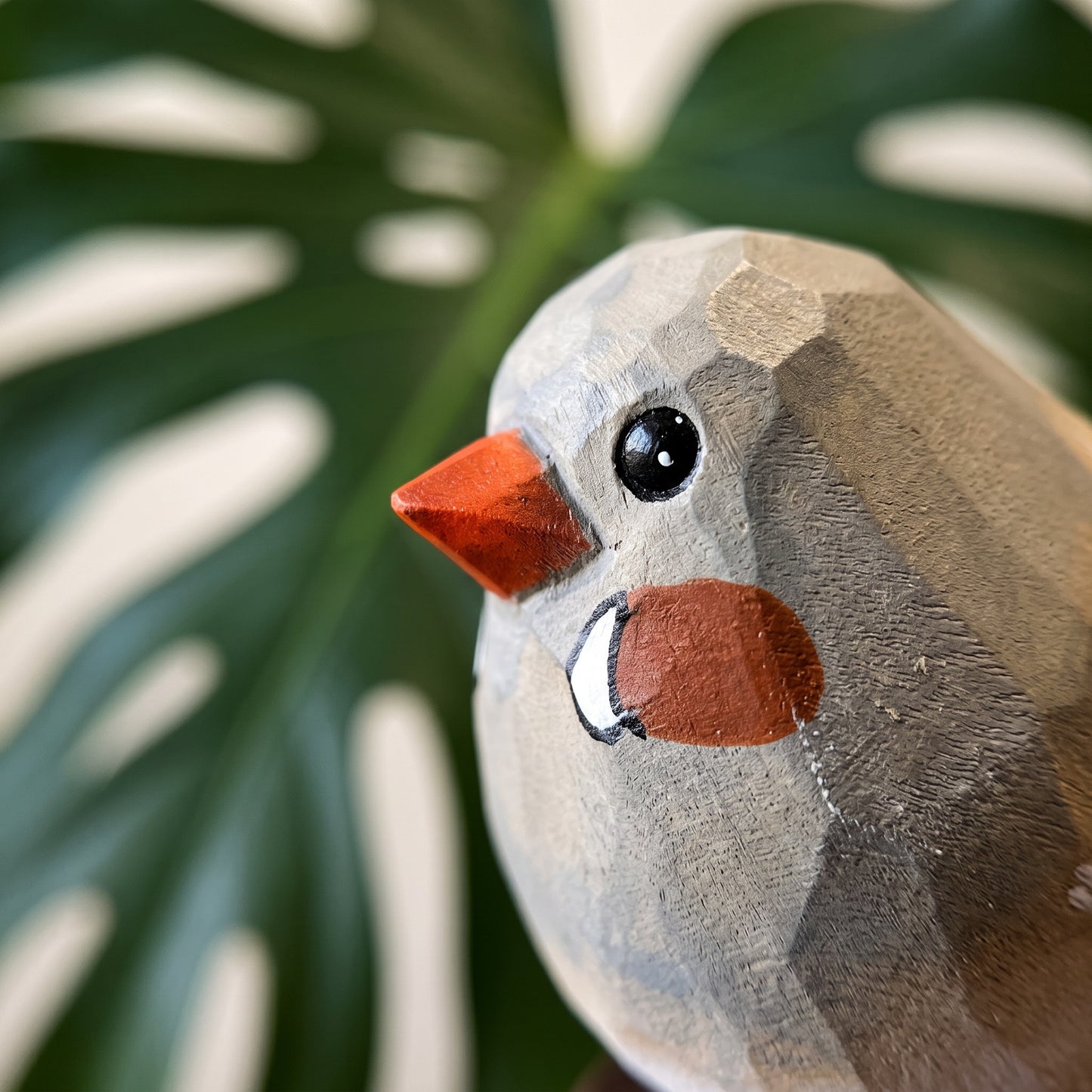 A close-up of a handcrafted wooden Zebra Finch figurine with geometric facets, featuring a gray-brown body, a bright orange beak, and a reddish-orange chest patch, set against a blurred green monstera leaf background.