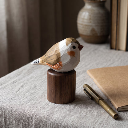 A handcrafted wooden Zebra Finch figurine with geometric facets, perched on a dark wood cylinder base. The bird features natural wood tones with hand-painted white, brown, and orange details.