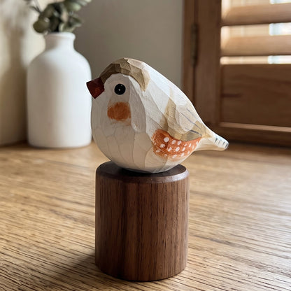 A small carved wooden Zebra Finch figurine with a dark brown cylindrical base, displayed on a wooden table in a softly lit room with books and a vase in the background.