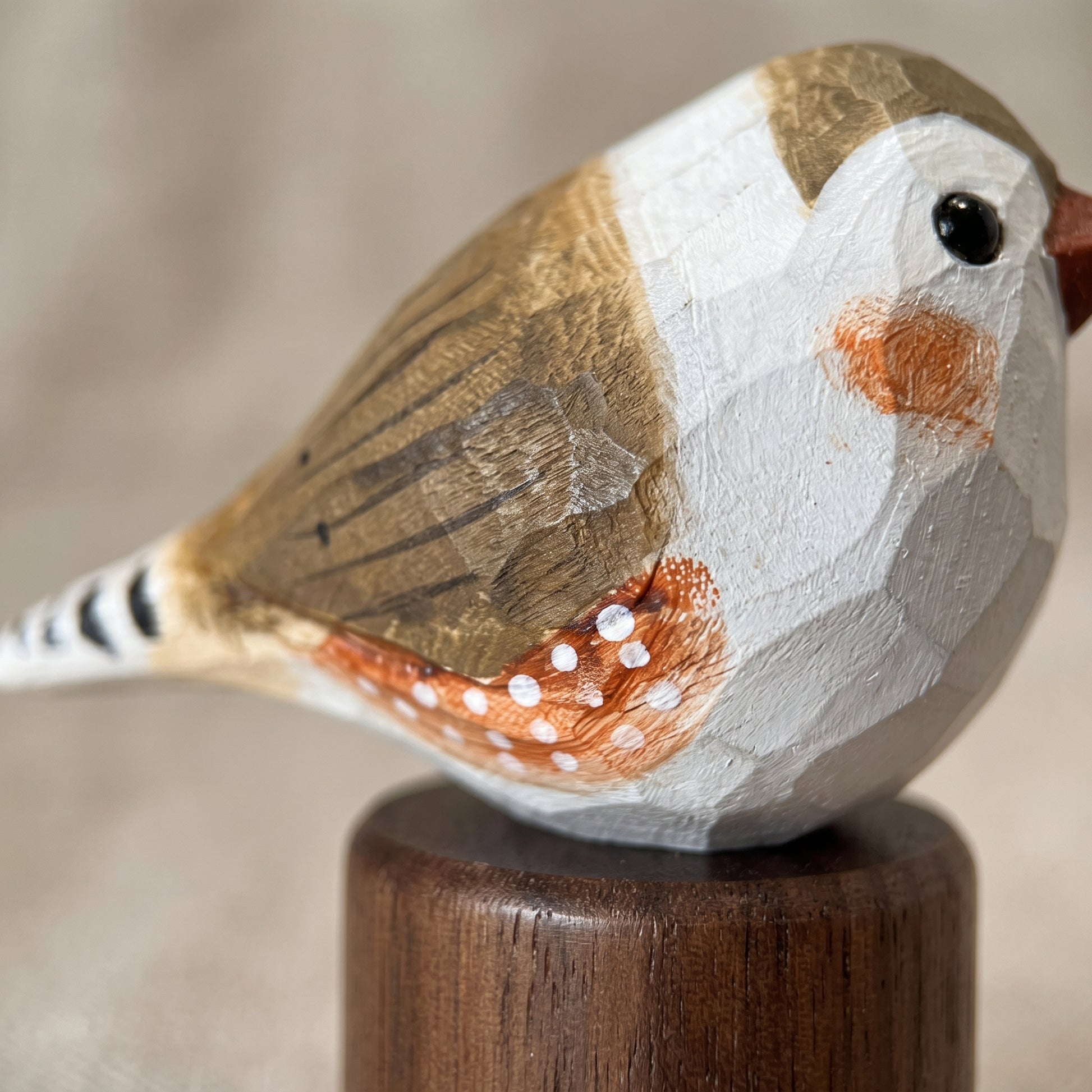 A close-up of a hand-carved wooden Zebra Finch figurine with painted brown, white, and orange details, mounted on a dark wooden stand against a soft beige background.