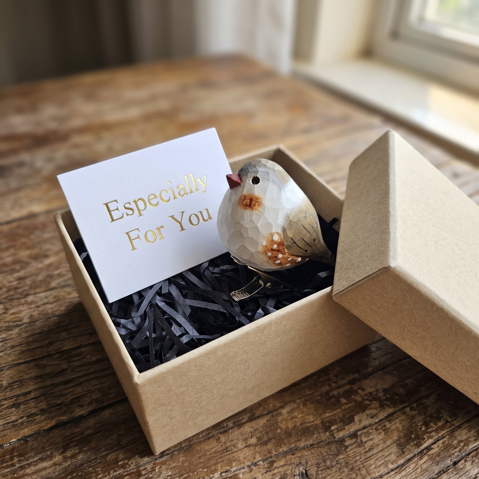 A hand-carved wooden zebra finch ornament with orange cheek patches, a black-and-white barred chest, and speckled sides perches inside an open kraft gift box filled with black shredded paper next to a white card reading "Especially For You".