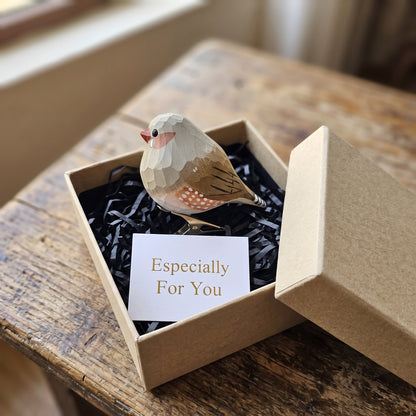 A hand-carved wooden zebra finch ornament with orange cheek patches and speckled sides perches inside an open kraft gift box filled with black shredded paper, alongside a white card that reads "Especially For You".